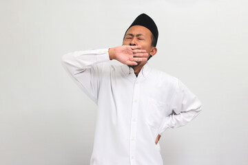 a Tired young Indonesian man wearing a songkok or peci or kopiah, yawns, covering his mouth with...