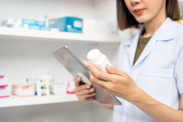 Beautiful asian woman pharmacist checks inventory of medicine in pharmacy drugstore. Professional Female Pharmacist wearing uniform standing near drugs shelves working with tablet.
