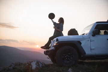 Girl traveler in hat sits with mug on hood of car against backdrop of sunset in mountains.