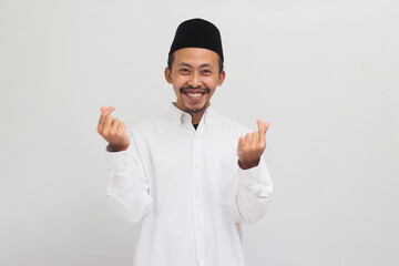 Smiling Young Indonesian man, wearing a songkok, peci, or kopiah, makes a mini heart hand sign with his finger, isolated on white background - obrazy, fototapety, plakaty