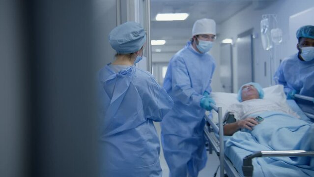 Professional surgeons transport old patient from successful operation to his hospital room down clinic hallway. Medics open doors from surgery for colleagues. Senior man lies on gurney under drip.