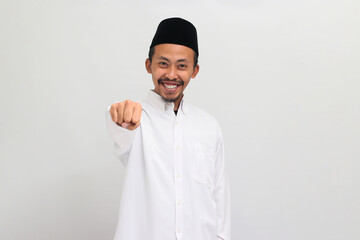 Confident young Indonesian man, wearing a songkok, peci, or kopiah, making fist bump gesture towards camera, isolated on a white background