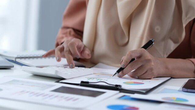 Motivated Muslim businesswoman wearing hijab working on real estate project analyzing document data graph discussing presentation work income calculation tax to a company in the office.