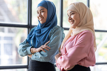 Portrait of successful and happy Two Young muslim women in hijab, office worker smiling and looking at camera with crossed arms, working inside modern office.