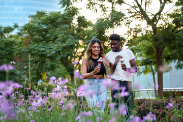 A beaming Afro-American man and a Latin woman, both in casual urban attire, savoring ice cream while strolling in a lush, flowery park at sunset.
