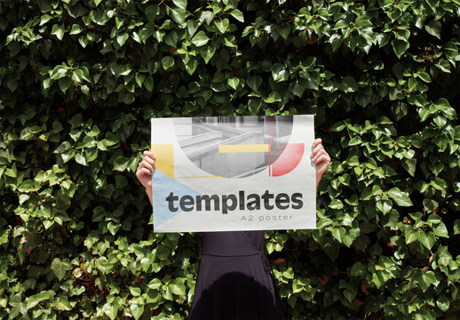 Mockup of person holding customizable A2 horizontal poster in front of face by hedge