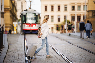 Travel carefully and safety with insurance and avoid accidents. Woman tourist with suitcase walks across city street while tram arrives - obrazy, fototapety, plakaty