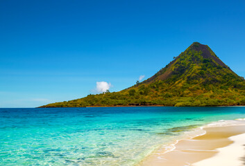 Remote tropical island with a volcanic peak, surrounded by crystal-clear waters and white sandy beaches, under a brilliant blue sky.