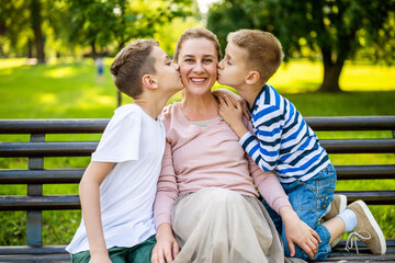 Happy mother is sitting with her sons on bench in park. They are having fun together. Boys are kissing their mother.