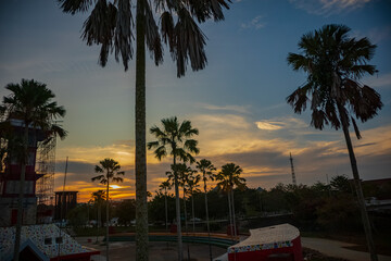 Obraz premium The sun sets in the Tenggarong city park with a building surrounded by palm trees.
