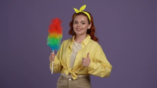 Woman in casual clothing posing at the camera, holding colorful pipidaster brush in hands, shows thumbs up. Isolated on purple background.