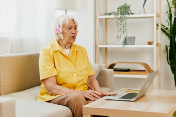 An elderly woman wearing headphones with a laptop sitting on the couch at home and working in a...