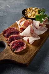Close-up vertical side angle, magnifying the textures and essence of a meat plate: roast beef, turkey pastrami, and chicken pâté on a rich wooden surface and gray concrete