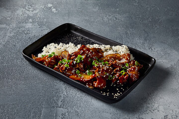 Horizontal side-view of the Kung Pao chicken, emphasizing the vibrant colors and texture on the black plate. The sweet-sour sauce glistens against the serene gray backdrop