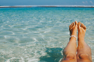 Beautiful women's legs with a bracelet against the background of clear azure ocean water. Stylish travel and tourism banner with blank space for text