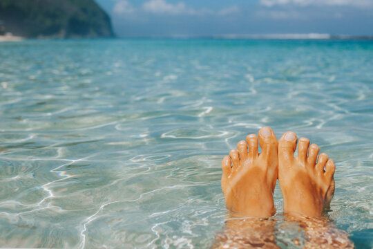 Women's tanned feet with a natural pedicure against the background of clear clear water and the tropics. Stylish banner of travel and leisure.