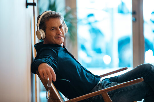 Smiling mature man wearing wireless headphones sitting in armchair