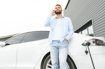 Happy man using smart phone and charging car at vehicle charging station