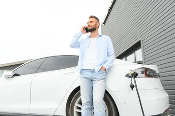 Casual man with smartphone near electric car waiting for the finish of the battery charging process