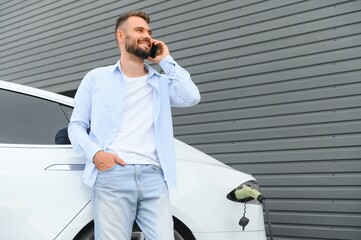 Casual man with smartphone near electric car waiting for the finish of the battery charging process