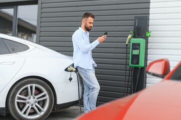 Happy man using smart phone and charging car at vehicle charging station