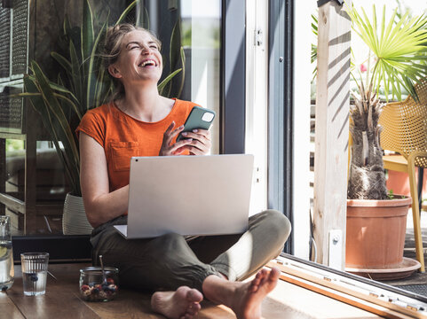Portrait of woman sitting by open balcony laughing with smart phone in hands