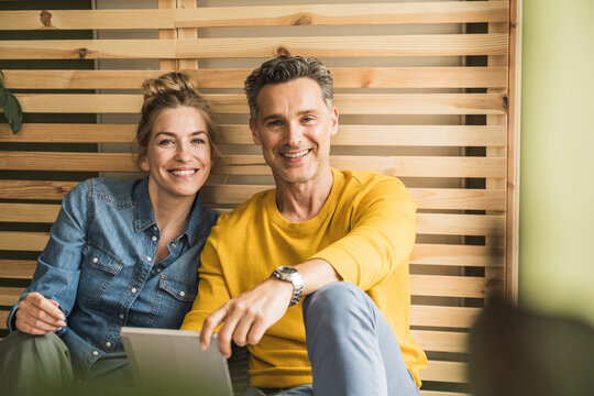 Portrait of couple sitting together in front of wooden bed frame leaning on wall - Powered by Adobe