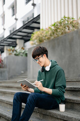 Handsome and talented young Asian man student working on his project assignment, using laptop computer and tablet to search an online informations..