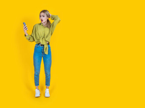 Confused Shocked Young Woman, Full Body Front View Beautiful Blonde Confused Shocked Young Woman. Puzzled Stressed Sad Caucasian Lady Holding Using Mobile Cell Phone Isolated On Yellow Background.