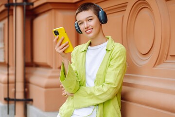 Summer portrait of a young woman with a short haircut with a phone and wireless headphones, listens to music and walks along the streets of a sunny city. Woman wearing headphones posing outdoors.
