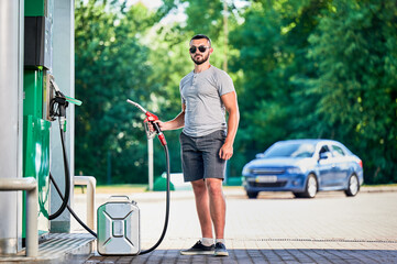 Fototapeta na wymiar Handsome driver refueling canister with gasoline at modern gas station. Portrait of man filling cistern with fuel in case of unforeseen circumstances. Man with pump nozzle on background of his car.