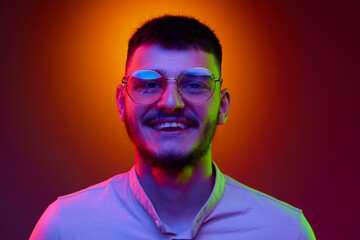 Fototapeta premium Happy smiling young guy, student in glasses against burgundy background in mixed color light, filter.