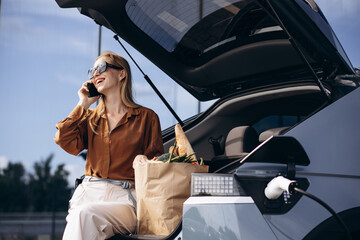 Woman with food shopping bags charging electric car and using mobile phone