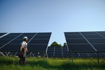Competent energy engineer in grey overalls and white helmet checking solar panels while walking on...