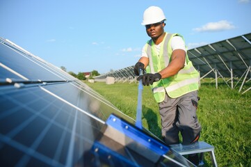 African-American worker in gray overalls and a white hard hat works in a field of solar panels. Solar renewable energy concept.