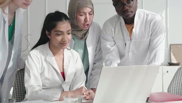 Happy multicultural international doctors interns working on laptop. Team of Medical Research Doctors Discuss Treatment, Medicine, Patients giving high five each other. Hospital Conference Meeting
