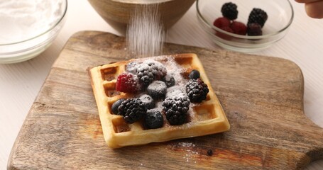 Powdered sugar is sprinkled on a Belgian waffle with blueberries, blackberries and raspberries. Breakfast preparation. Sweet food.