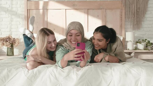 Diverse happy excited multiethnic young women friends people laughing talking together, laying on a bed and making selfie photo, celebrating friendship, capture moments, memories. Sisters
