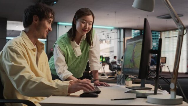 Caucasian Male Game Designer Using Desktop Computer With 3D modelling Software And Talking to Asian Female Art Director in Game Development Studio Office. Diverse Colleagues Discussing New RPG Game.