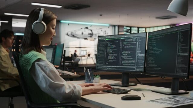 Female Asian Game Programmer Coding On Desktop Computer In Game Design Studio Diverse Office. Woman Wearing Headphones, Writes Lines Of Code. Gameplay Engineer of New Immersive 3D Survival Video Game.