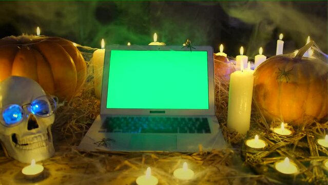 Laptop with chromakey green screen in fog among burning candles on floor. Traditional pumpkins and spiders, pentagram, creepy autumn Happy Halloween background, moving away tracking shot close up.