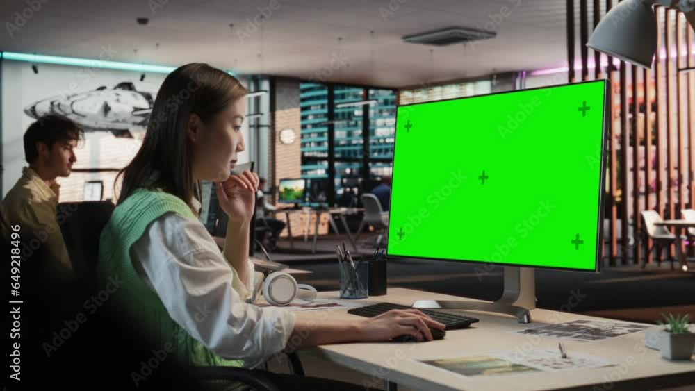 Poster Creative Asian Woman Sitting at Her Desk Using Desktop Computer with Mock-up Green Screen Chromakey. Female Level Designer Working in Game Design Studio Diverse Office, Creating Immersive Gameplay.
