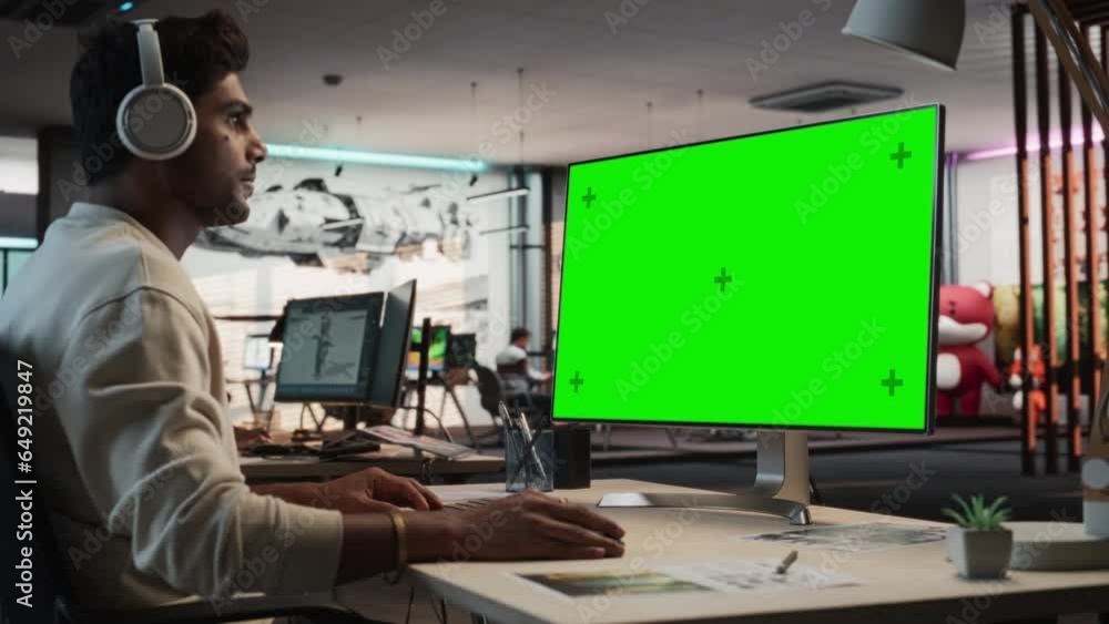 Leinwandbilder Portrait of Creative Indian Man Sitting at a Desk Using Desktop Computer with Mock-up Green Screen Chromakey. Male Level Designer Working in Game Design Startup Office, Creating Immersive Gameplay.
