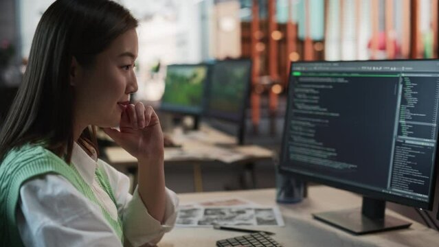 Female Asian Game Programmer Coding On Desktop Computer In Game Development Studio Diverse Office. Focused Woman Writes Lines Of Code, Does AI Programming For New Immersive 3D Survival Video Game.