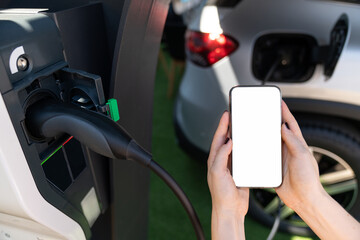 Hands with smartphone and charging plug on a background of electric car . White screen, you can add...