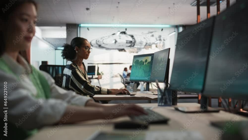 Aufkleber Focus Shifting From Female Asian Game Programmer Using Desktop Computer to Black Woman At a Desk Working On Immersive Game World In 3D modelling Software on PC. People Working In Game Design Studio.