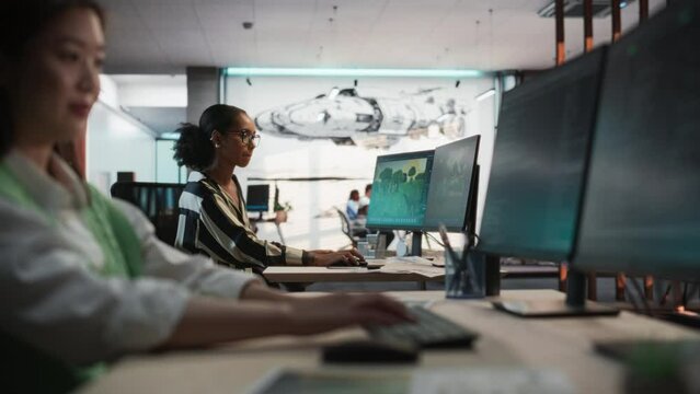Focus Shifting From Female Asian Game Programmer Using Desktop Computer to Black Woman At a Desk Working On Immersive Game World In 3D modelling Software on PC. People Working In Game Design Studio.