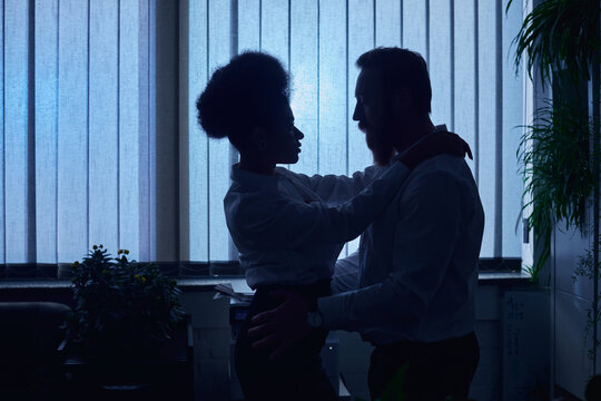 dark silhouettes of multiethnic couple embracing in office at night, romantic encounter at work