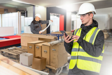 Man in carpentry shop. Worker is holding phone. Pallets with boxes in production room. Employee of carpentry shop writes SMS. Man with tablet computer. Two workers inside carpentry factory