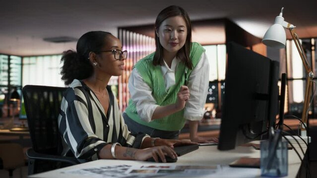 Black Female Game Designer Using Desktop Computer With Animation Software And Talking to Asian Female Project Manager in Game Development Studio Office. Diverse Colleagues Discussing New RPG Game.
